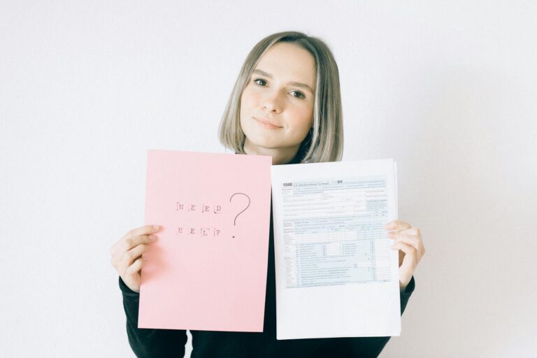 A woman in a black shirt holds tax forms and a 'Need Help?' sign, indoors.